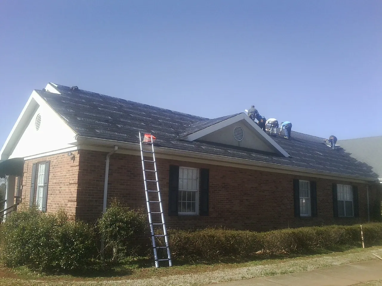 Crew tearing shingles off a brick ranch roof with a ladder set — Emergency Roof Repair work by our crew in Bay Saint Louis