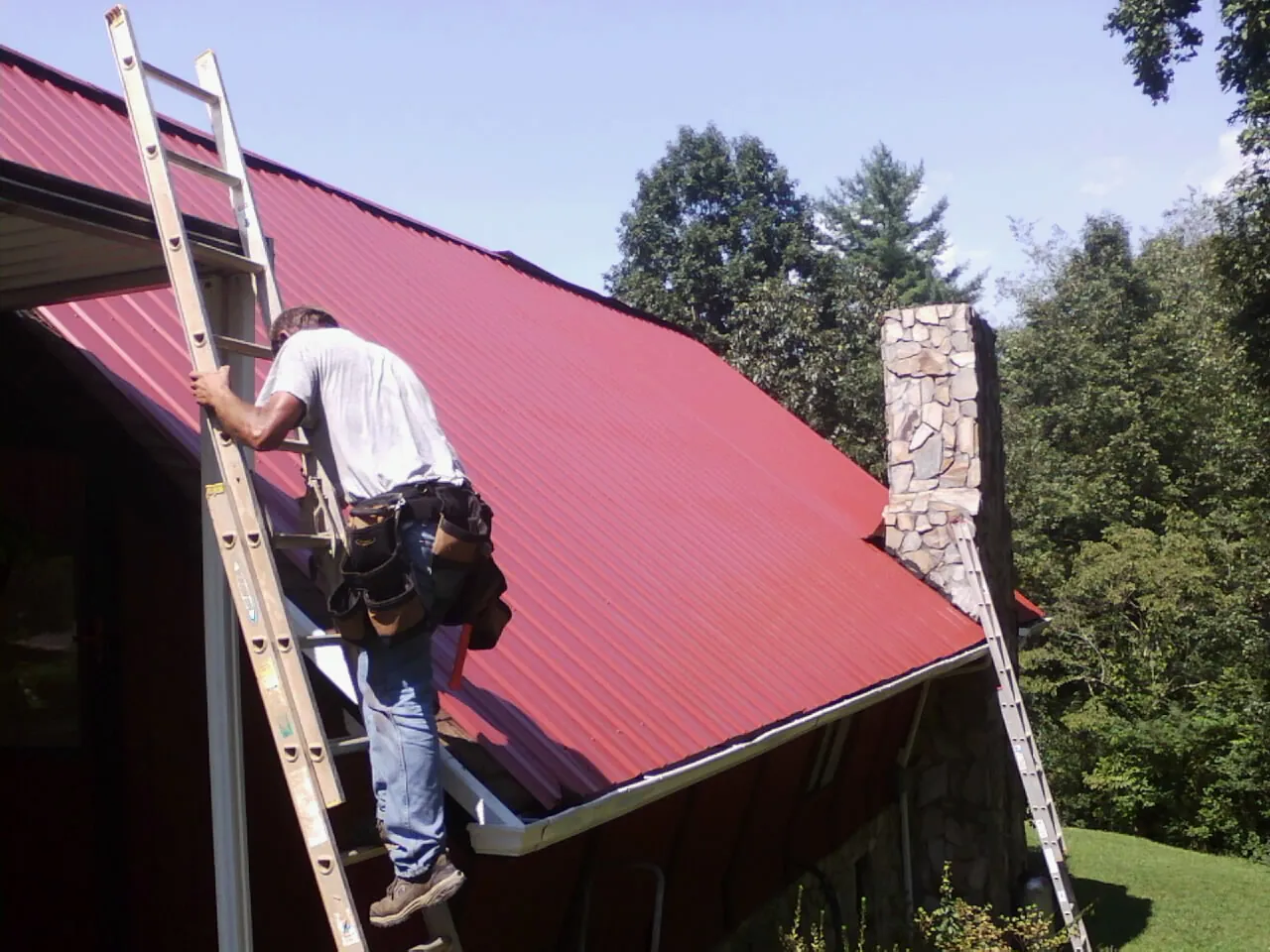 Roofer on a ladder beside a red metal roof and stone chimney — Soffit & Fascia Repair by Roofing Brothers in Bay Saint Louis